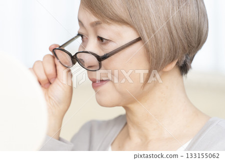Senior woman wearing reading glasses and looking at a hand mirror in the living room Senior woman wearing reading glasses and looking at a hand mirror in the living room 133155062