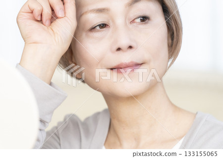 Senior woman wearing reading glasses and looking at a hand mirror in the living room 133155065