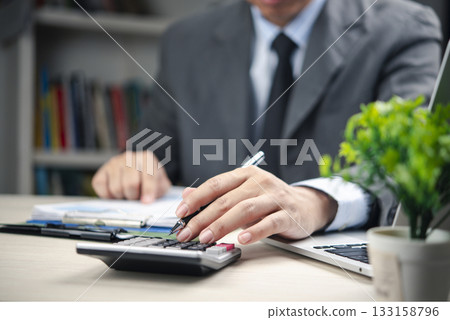 Businessman using calculator and reviewing financial report. Concept of accounting, bookkeeping payroll and budget tax., business finance, and professional financial planning at office desk. Businessman using calculator and reviewing financial report. Concept of accounting, bookkeeping payroll and budget tax., business finance, and professional financial planning at office desk. 133158796
