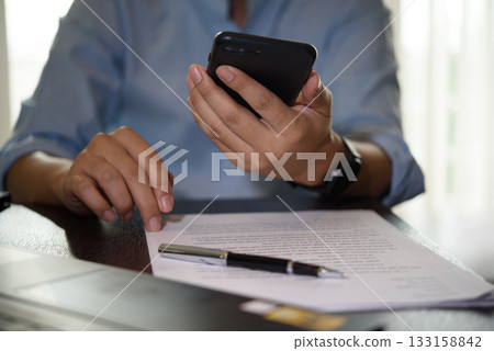 Hand holding smartphone while reviewing business document. Mobile communication, digital signature, and financial contract approval in remote work environment. 133158842