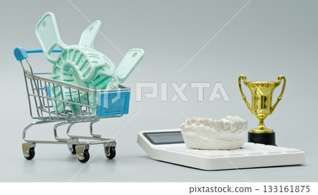 Mini shopping cart filled with dental impression trays beside a jaw model, calculator, and trophy, symbolizing cost, quality, and dental excellence. Mini shopping cart filled with dental impression trays beside a jaw model, calculator, and trophy, symbolizing cost, quality, and dental excellence. 133161875