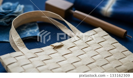 Handwoven cream colored wicker basket with double handles and herringbone pattern displayed on blue fabric surface with leather crafting tools in background 133162309
