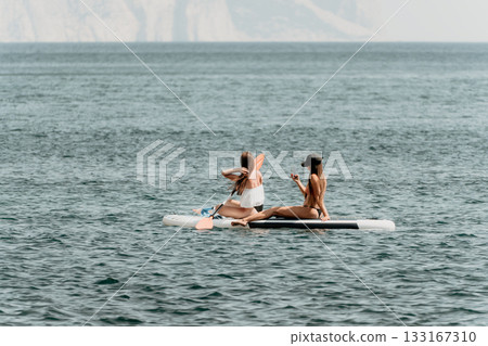 Two women floating on a surfboard in the ocean. One of them is wearing a white shirt and the other is wearing a black bikini. Two women floating on a surfboard in the ocean. One of them is wearing a white shirt and the other is wearing a black bikini. 133167310