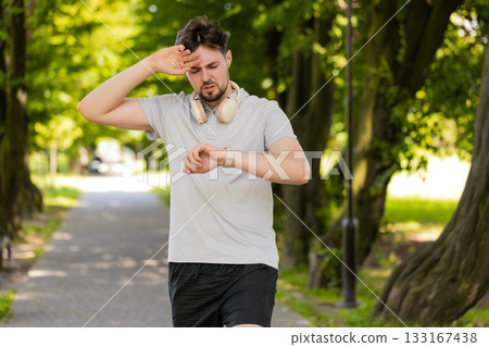 Active young man jogger running in public park and looking at smartwatch, getting tired relaxing 133167438