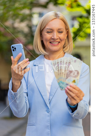 Caucasian mature businesswoman on city street showing earnings cash money with smartphone in hands Caucasian mature businesswoman on city street showing earnings cash money with smartphone in hands 133167445
