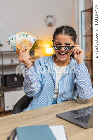Young woman freelancer putting on party glasses holding fan of cash sitting at home office table Young woman freelancer putting on party glasses holding fan of cash sitting at home office table 133167477