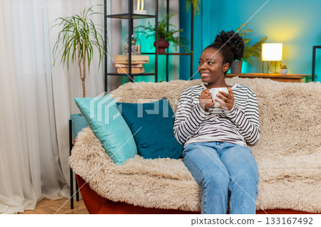 African American young woman relaxing at home on couch, holding hot tea cup, dreaming, smiling African American young woman relaxing at home on couch, holding hot tea cup, dreaming, smiling 133167492