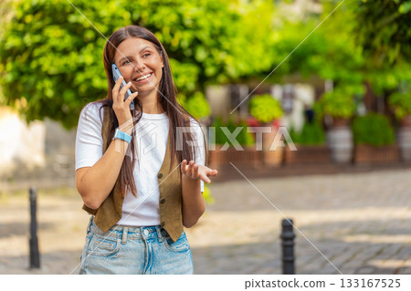 Happy young Caucasian woman in casual talking gossip on smartphone while walking on city street 133167525