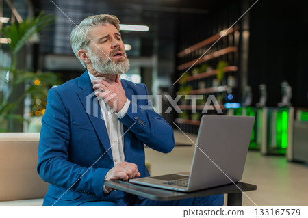 Sick businessman talking on video call having sore throat, coughs sitting with laptop in lobby Sick businessman talking on video call having sore throat, coughs sitting with laptop in lobby 133167579