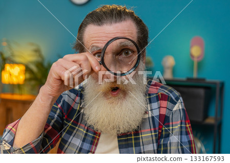 Investigator researcher scientist senior man working at home holding magnifying glass near face Investigator researcher scientist senior man working at home holding magnifying glass near face 133167593