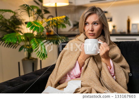Adult woman sick with sore throat in blanket holds tea takes sips wipes nose with tissues at night Adult woman sick with sore throat in blanket holds tea takes sips wipes nose with tissues at night 133167603