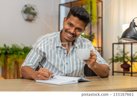 Indian man writes goals in notebook sips coffee ticks boxes smiles and plans day focused at desk Indian man writes goals in notebook sips coffee ticks boxes smiles and plans day focused at desk 133167661