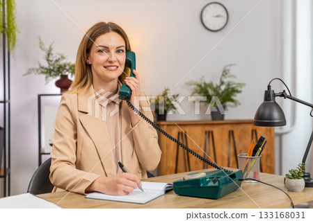 Businesswoman calls client on retro phone spins rotary dial takes notes agreement formal tone smile Businesswoman calls client on retro phone spins rotary dial takes notes agreement formal tone smile 133168031