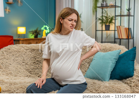 Pregnant woman holds lower back feels severe pain in spine near sofa handles daily pain needs relief Pregnant woman holds lower back feels severe pain in spine near sofa handles daily pain needs relief 133168047