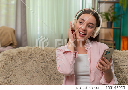 Young woman dancing on sofa wearing wireless headphones enjoying favorite music with joyful energy Young woman dancing on sofa wearing wireless headphones enjoying favorite music with joyful energy 133168055