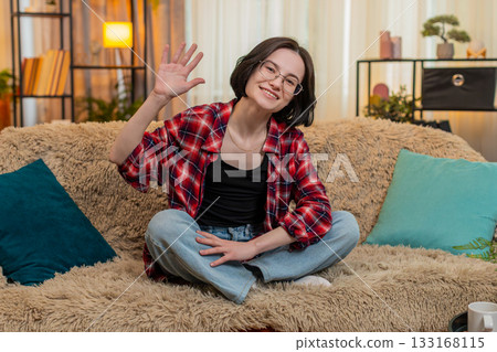 Young woman smiling and waving hand at home, greeting someone with friendly and warm expression Young woman smiling and waving hand at home, greeting someone with friendly and warm expression 133168115