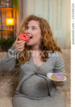 Pregnant young woman eating donuts on home sofa laughing with joyful expression enjoying sweet treat Pregnant young woman eating donuts on home sofa laughing with joyful expression enjoying sweet treat 133168120