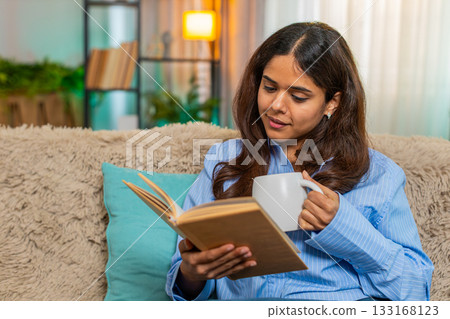 Indian woman drinking coffee and reading book at home enjoying quiet peaceful start of cozy morning Indian woman drinking coffee and reading book at home enjoying quiet peaceful start of cozy morning 133168123
