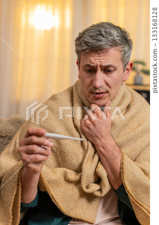 Young man checks body temperature with thermometer looks worried about possible illness and fever Young man checks body temperature with thermometer looks worried about possible illness and fever 133168128