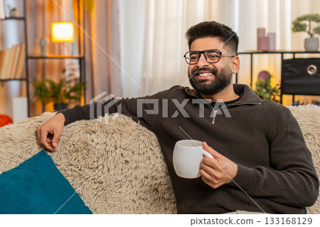 Indian man at home sofa enjoys tea or coffee after long day, smiling and relaxing in peaceful mood Indian man at home sofa enjoys tea or coffee after long day, smiling and relaxing in peaceful mood 133168129