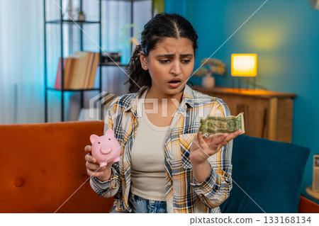 Woman sadly looking at piggy bank with one dollar inside financial difficulties limited savings 133168134