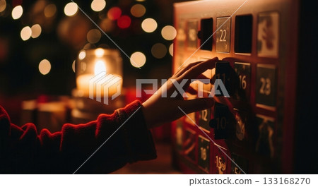 A child's hand opens a wooden advent calendar, bathed in the warm glow of a candle and festive lights. This cozy scene captures the magic and anticipation of the holiday season. 133168270