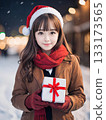 Smiling young woman holding a gift on a snowy night with sparkling Christmas lights 133173565