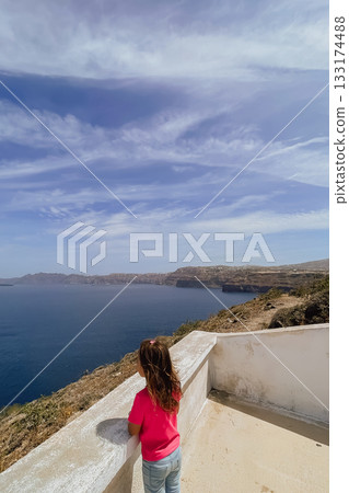 Child enjoying the stunning cliffside view over the water on a sunny day in the picturesque Greek island of Santorini 133174488