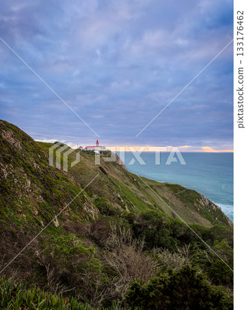Scenic ocean view of Cabo da Roca or Cape Roca, Portugal, lighthouse on high rugged cliffs at sunset over horizon, ocean waves and surrounding greenery of landscape. Famous travel destination 133176462