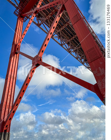 Upshot view of tower and main girder of the famous red April 25th or 25 de Abril bridge in Lisbon, Portugal, against blue sky with fluffy white clouds on sunny day. Tourist attraction, travel 133176469