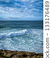 Clear ocean waves roll in with foamy edge, rocky shoreline in foreground with scarce greenery, heavy clouds in blue sky on sunny day, distant horizon. Atlantic ocean landscape, stormy weather forecast 133176489