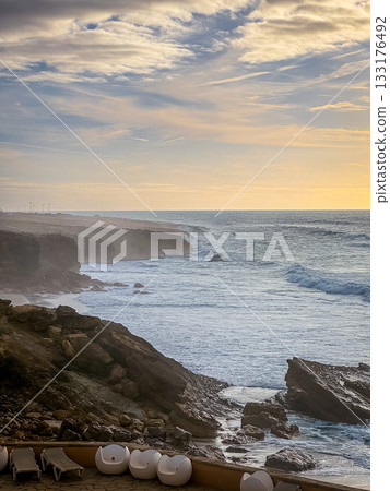 Scenic view of turbulent ocean waves hitting rugged cliffs as sun sets in background. White lounge chairs are scattered on terrace balcony, serene atmosphere. Ocean view, sundown sky with clouds.  133176492