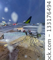 Snowy airport scene shot through airplane window covered in melting snowflakes and deicing fluid. Ground crew members working, another aircraft waiting in distance. Winter storm covering airport 133176494
