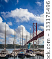 Several sailing boats anchored below famous red April 25th or 25 de Abril bridge in Lisbon, Portugal in river Tejo or Tagus estuary. Fluffy clouds in blue sky on sunny day, monument to Christ the King 133176496