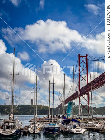 Several sailing boats anchored below famous red April 25th or 25 de Abril bridge in Lisbon, Portugal in river Tejo or Tagus estuary. Fluffy clouds in blue sky on sunny day, monument to Christ the King 133176496
