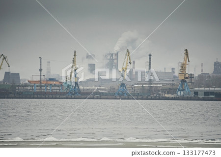 Industrial landscape, a factory with pipes and cranes in the port against a gray foggy sky Industrial landscape, a factory with pipes and cranes in the port against a gray foggy sky 133177473
