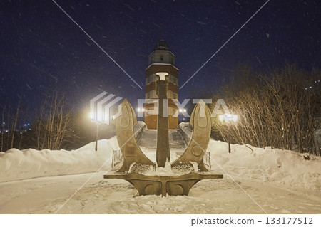 Large anchor and lighthouse in the city against the background of the night sky in snowfall in winter 133177512