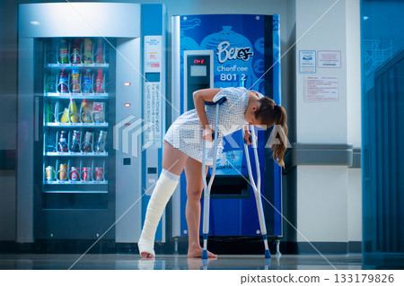 Injured woman struggles at hospital vending machine. A woman using crutches stands in front of a vending machine in a hospital setting.  133179826