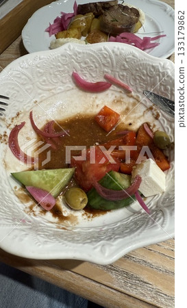Half-eaten greek salad on ornate white plate with cucumbers, tomatoes, feta, and olives 133179862
