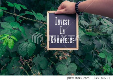 Hand holding a framed sign with the phrase Invest In Knowledge among lush green foliage Hand holding a framed sign with the phrase Invest In Knowledge among lush green foliage 133180727