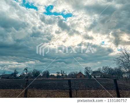 Dramatic cloudy sky over rural landscape with fields and distant houses under natural light 133180757