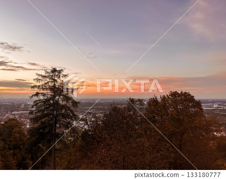 Scenic sunset view from a hilltop with vibrant colors illuminating the sky, silhouetting trees and showcasing a panoramic landscape below 133180777