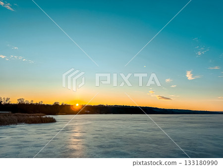 Serene winter landscape at sunset with frozen lake reflecting vibrant colors and distant trees, creating a tranquil and picturesque atmosphere 133180990