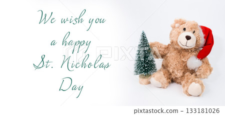 Plush teddy bear wearing a red hat holding a small green Christmas tree with festive greeting 133181026