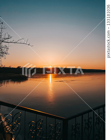 Serene sunset over tranquil lake with reflections on water, surrounded by trees and a decorative railing, creating a peaceful natural atmosphere Serene sunset over tranquil lake with reflections on water, surrounded by trees and a decorative railing, creating a peaceful natural atmosphere 133181036
