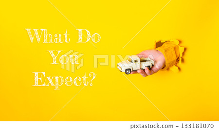 Hand holding a toy truck emerging from a bright yellow background with text what do you expect 133181070