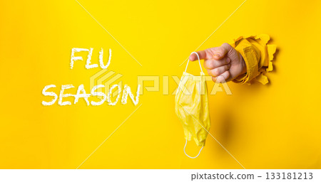 Hand holding yellow medical mask through torn paper, emphasizing flu season awareness and prevention Hand holding yellow medical mask through torn paper, emphasizing flu season awareness and prevention 133181213