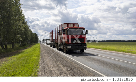 Logging truck with a loader drives along the road. A front view of a long, empty red industrial logging trailer moving along the highway against a blue summer sky. 133181930