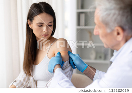 Doctor applying adhesive bandage on female patients arm after vaccination in clinic 133182019