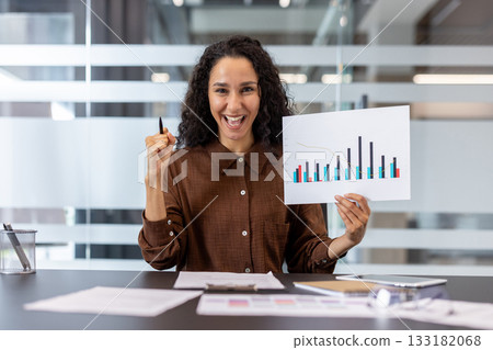 Mixed race businesswoman celebrating business success and achievement, holding a chart with increasing bars and a pen, expressing excitement while sitting at her office desk 133182068